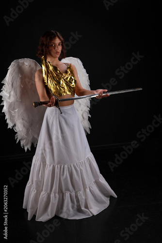 Full length cinematic portrait of a beautiful woman in a golden corset and white angel wings holding a medieval sword on a black background