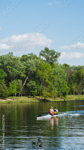 Rear view of two rowers training in a double scull boat on a calm river with lush green forest on the background.
