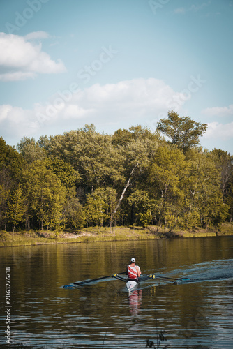 Back view of a male rower in a sculling boat training on a calm river with green trees and blue sky on the background.