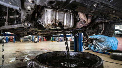 A mechanic changes oil under a car. Dark oil drains into a black container. The mechanic wears blue gloves and holds a tool. The garage is well-lit with tools in the background.