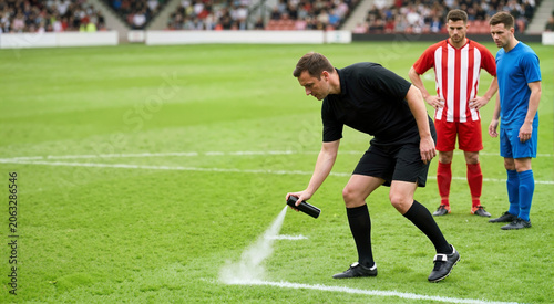 Football referee spraying white vanishing foam on grass pitch for free kick. Soccer players watching in stadium