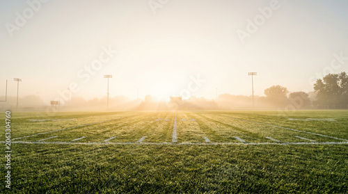 A scenic view of an empty American football field at sunrise with golden sun rays piercing through heavy morning fog over dew-covered green grass and white yard lines in a stadium setting