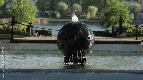 dark, spherical stone fountain spraying water in a sunny garden. The water droplets sparkle in the sunlight, framed by a beautiful patch of blooming yellow daffodils.