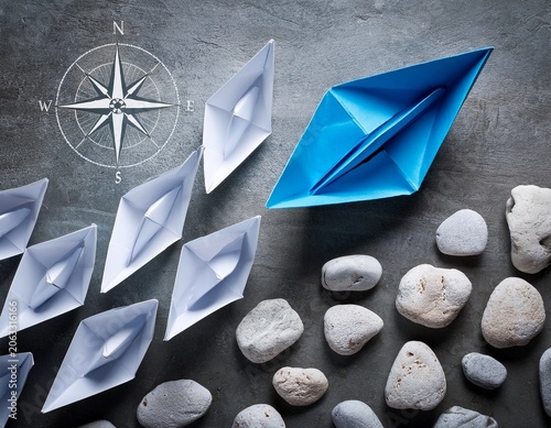 blue paper boat with compass rose leading a fleet of small white boats away from rocks on grey concrete leadership concept