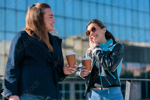 Two friends laugh and chat while enjoying takeaway coffee outside a modern building during a sunny day