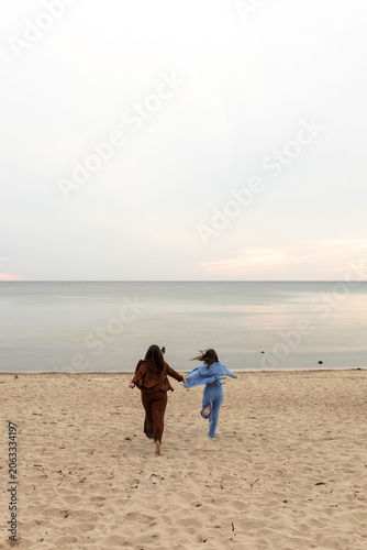 Two girlfriends walk hand in hand along a sandy beach towards the sea.
