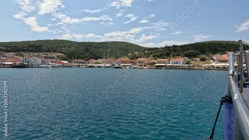Boat approaching a picturesque Greek coastal village on a sunny day in the Ionian Sea. Traditional waterfront houses, turquoise water, small harbor and Mediterranean island atmosphere create a scenic 
