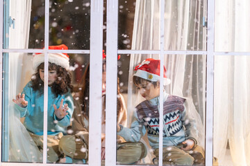 Excited children in Santa hats open Christmas presents together by a decorated tree and cozy fireplace at home.
