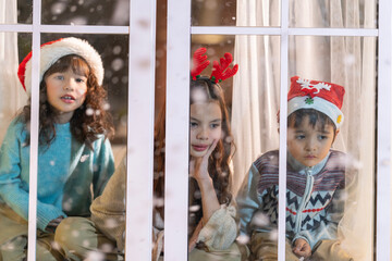 Excited children in Santa hats open Christmas presents together by a decorated tree and cozy fireplace at home.