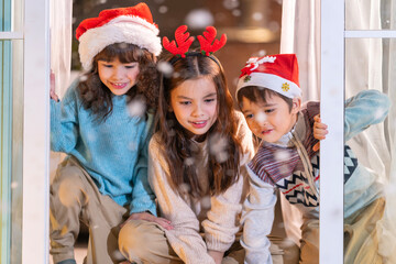 Excited children in Santa hats open Christmas presents together by a decorated tree and cozy fireplace at home.