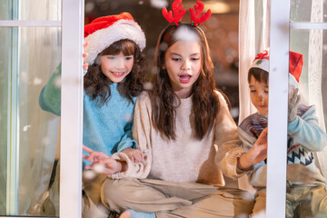 Excited children in Santa hats open Christmas presents together by a decorated tree and cozy fireplace at home.