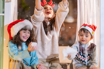 Excited children in Santa hats open Christmas presents together by a decorated tree and cozy fireplace at home.