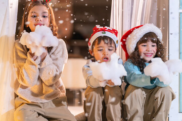 Excited children in Santa hats open Christmas presents together by a decorated tree and cozy fireplace at home.