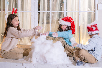 Children in Santa hats and reindeer antlers gather by a glowing decorated Christmas tree opening gifts at home.