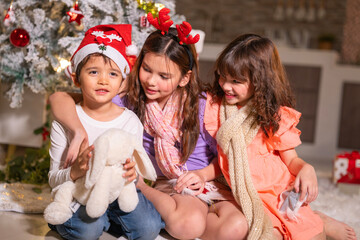 Children in Santa hats and reindeer antlers gather by a glowing decorated Christmas tree opening gifts at home.