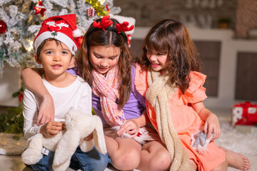 Children in Santa hats and reindeer antlers gather by a glowing decorated Christmas tree opening gifts at home.