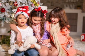 Children in Santa hats and reindeer antlers gather by a glowing decorated Christmas tree opening gifts at home.