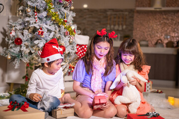 Children in Santa hats and reindeer antlers gather by a glowing decorated Christmas tree opening gifts at home.