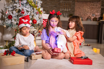 Children in Santa hats and reindeer antlers gather by a glowing decorated Christmas tree opening gifts at home.