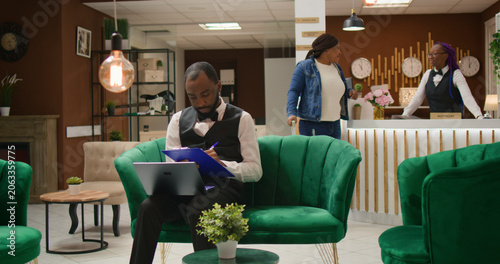 Bild auf Leinwand African American porter manages bookings before arrival of guests to ensure pleasant stay during vacation, hotel concierge man handling lodging accommodation on laptop, lounge area