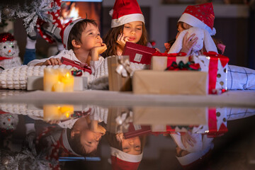 Excited children in Santa hats open Christmas presents together by a decorated tree and cozy fireplace at home.