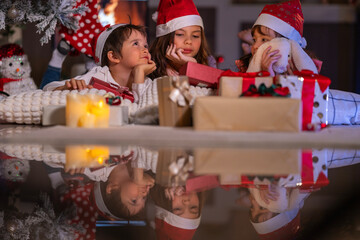 Excited children in Santa hats open Christmas presents together by a decorated tree and cozy fireplace at home.