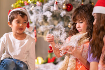Children in Santa hats and reindeer antlers gather by a glowing decorated Christmas tree opening gifts at home.