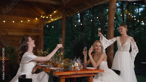 Three happy brides in white wedding dresses toasting with wine glasses at wooden table decorated with wildflowers under string lights in outdoor pavilion surrounded by forest