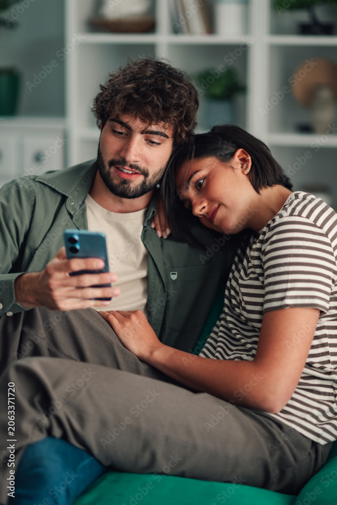 Obraz premium Young couple relaxing at home watching smartphone together, sharing social media content and digital connection