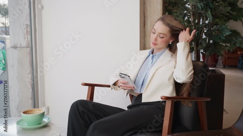 Woman sits in a cafe dressed in a blazer. She uses her phone while resting a cup of coffee on table nearby. She plays with her hair as she waits.