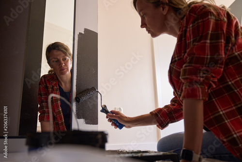 Woman applies dark paint to wall with roller beside mirror, with her reflection visible