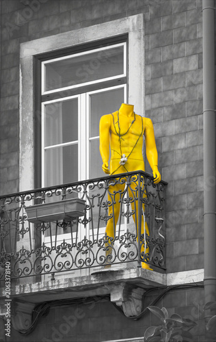 A bright yellow headless mannequin stands on an ornate balcony against a grayscale background. The contrast draws attention to the statue.