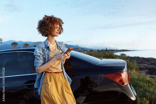A young woman with curly hair stands beside a dark car on a sunny coastal road, glancing at her phone while wearing a striped tee and denim shirt, embodying casual relaxed travel mood.