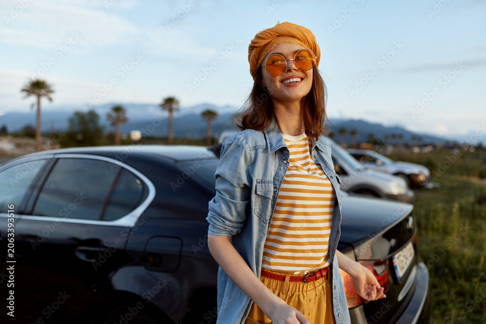 Fototapeta premium A cheerful woman in a striped shirt, denim jacket, orange beanie and sunglasses stands beside a parked car in a sunny outdoor setting. Lifestyle portrait of casual confidence.