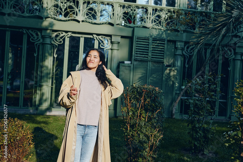 Canvas Print Woman in a long beige coat and jeans stands outdoors in sunlight, smiling, holding a drink, casual fashion scene in a residential garden setting