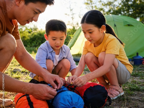 Asian father teaching children how to roll up sleeping bag at campsite for family outdoor education and summer camp concept