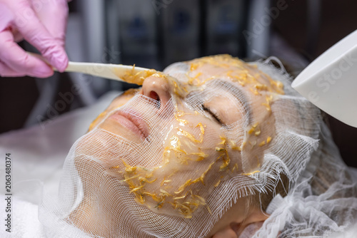 Professional cosmetologist in pink gloves applying alginate mask with calendula petals on woman's face at spa salon. Skin rejuvenation and hydration treatment.