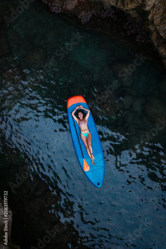 Drone shot of a woman relaxing on a paddleboard in a rocky sea canyon.