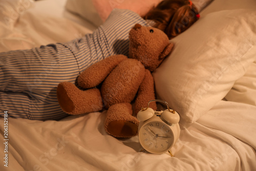 Alarm clock, teddy bear and sleeping little girl on bed