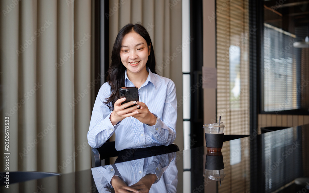 Fototapeta premium Young businesswoman smiling while using smartphone during a coffee break in a modern cafe workspace.