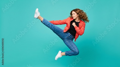 Woman jumping and performing a high kick wearing casual clothes against a turquoise background with energetic lifestyle strength movement