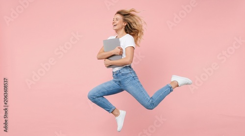 Woman running and jumping while carrying a laptop computer isolated on a pink background with computer technology lifestyle business