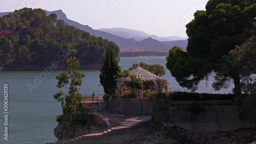 Drone footage panning sideways with telephoto compression past El Chorro's ornate white gazebo and stone promenade, layering jade inlet, pine island, and hazy mountain silhouettes behind.