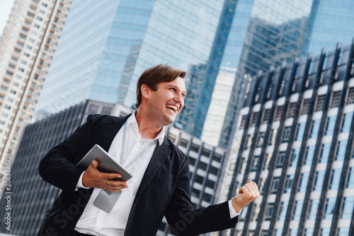 happy caucasian businessman or financial investor holding a tablet and celebrating success from stock market profit. Excited corporate executive standing outdoors against modern city office buildings