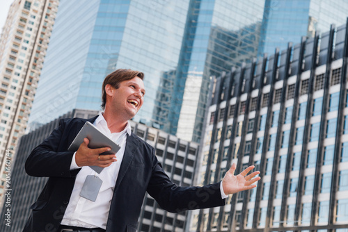happy caucasian businessman or financial investor holding a tablet and celebrating success from stock market profit. Excited corporate executive standing outdoors against modern city office buildings