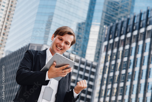 happy caucasian businessman or financial investor holding a tablet and celebrating success from stock market profit. Excited corporate executive standing outdoors against modern city office buildings