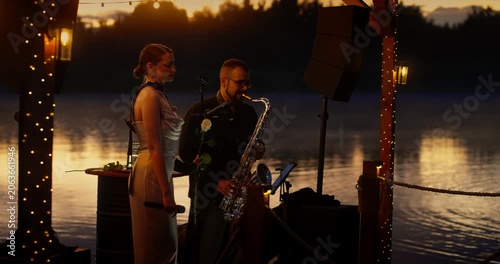 Female wedding singer and saxophonist performing on lakeside wooden stage at dusk