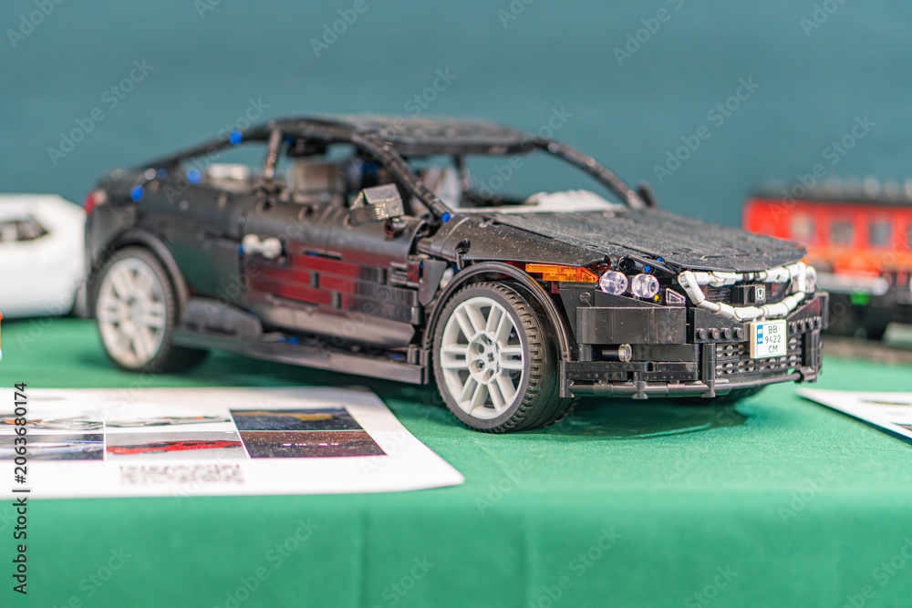 Naklejka premium Black and silver Lego replica of an Aston Martin DB9 toy car, elevated on a clear plastic stand against a green tablecloth Stacked white papers nearby - Kyiv, Ukraine - June 7, 2025