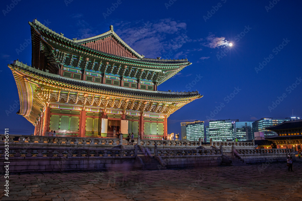 Fototapeta premium Stunning night view of Geunjeongjeon Hall at Gyeongbokgung Palace with full moon and modern city skyscrapers background in Seoul South Korea
