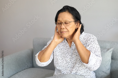 Asian Senior woman sits on a sofa at home with neck pain and muscle discomfort. She appears tired and stressed, showing health challenges and fatigue during her daily life.